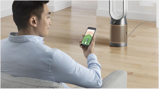 Un uomo seduto su un divano controlla un purificatore d’aria Dyson tramite un'app sul suo smartphone, in un ambiente domestico moderno con pavimento in legno chiaro.